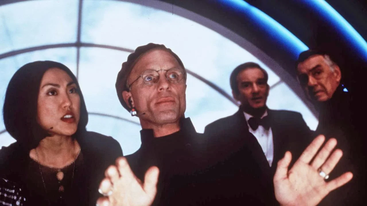 Christof raises both hands while directing from a control room filled with production staff and surveillance screens