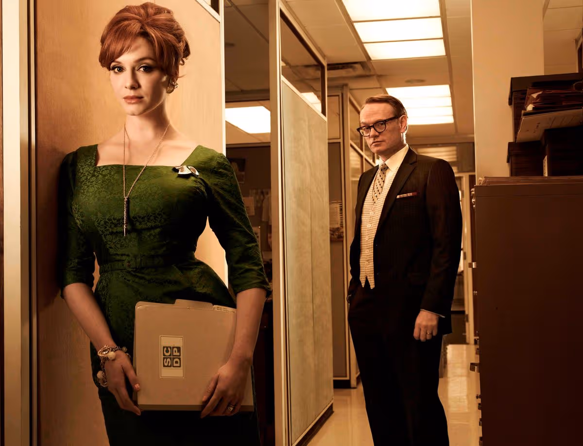 Joan Holloway stands in the foreground of the Sterling Cooper office while Lane Pryce watches from behind