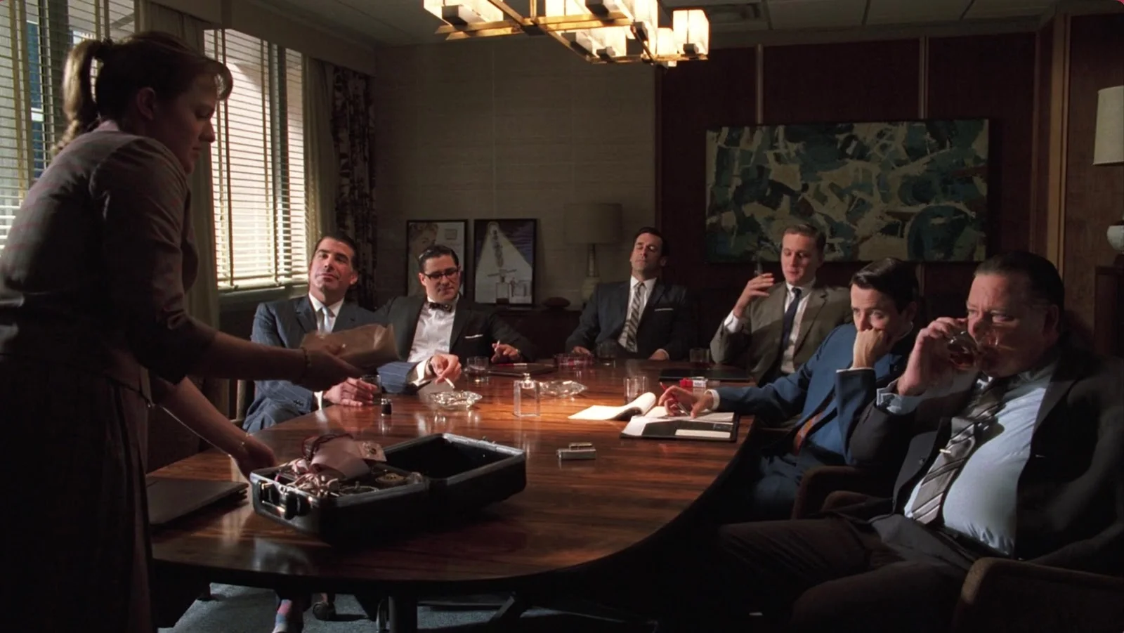 Sterling Cooper executives sit around a conference table while a woman serves drinks